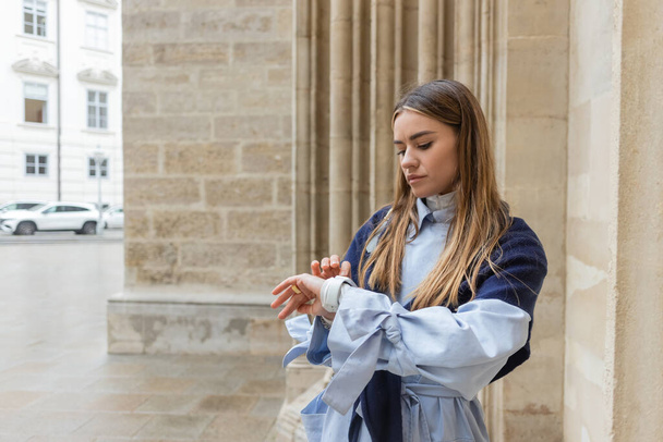 Junge Frau mit Schal auf blauem Trenchcoat checkt Zeit auf Smartwatch nahe historischem Gebäude in Wien  - Foto, Bild
