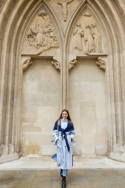 šťastná žena s šátkem na modrém plášti, stojící s rukama v kapsách poblíž historické budovy ve Vídni  - Fotografie, Obrázek