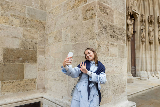 happy woman with scarf on top of blue trench coat having video call near historical building in Vienna  - Фото, зображення