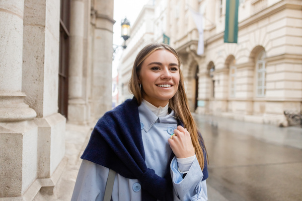 happy and stylish woman with scarf on top of blue trench coat smiling while looking away on street in Vienna - Fotó, kép