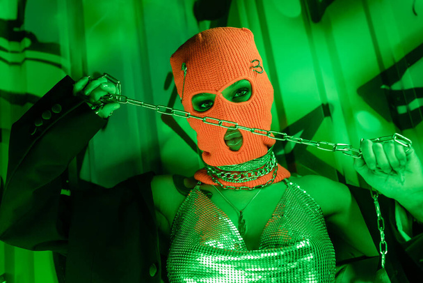 mujer provocativa en pasamontañas de punto y top plateado sosteniendo cadena metálica cerca de la pared con graffiti en iluminación verde - Foto, imagen