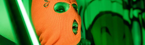 Parlak neon lambanın yanında örülmüş turuncu kar maskeli seksi bir kadın portresi ve grafitiyle yeşil bir duvar. - Fotoğraf, Görsel