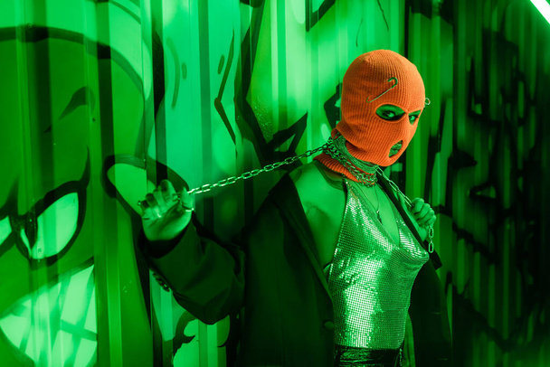 provocative woman in balaclava and sexy outfit posing with neck chain near wall with graffiti in green lighting - Φωτογραφία, εικόνα
