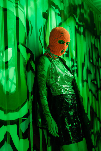szexi nő fekete bőr szoknya és ezüst tetején narancssárga maszk áll közel zöld fal graffiti - Fotó, kép