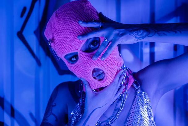 portrait of seductive woman in balaclava and silver top posing with hands near face at wall with graffiti in blue neon lighting - Photo, Image