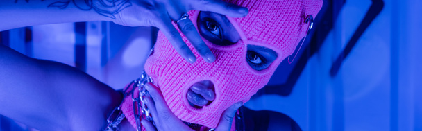 provocative woman in balaclava posing with hands near face at wall with graffiti in blue neon lighting, banner - Photo, Image
