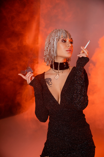 extravagante mulher tatuada em peruca de prata e vestido sexy posando com cigarro no fundo vermelho e laranja com fumaça - Foto, Imagem