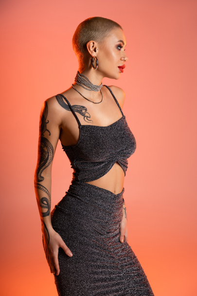 tattooed woman in grey lurex crop top and skirt looking away on coral pink background - Fotoğraf, Görsel