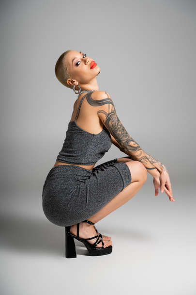 full length of tattooed and sexy woman in lurex clothes and sandals sitting on haunches and looking at camera on grey background - Fotografie, Obrázek