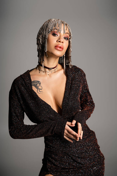 tattooed woman with sexy body posing in black lurex dress and jewelry wig isolated on grey - Zdjęcie, obraz