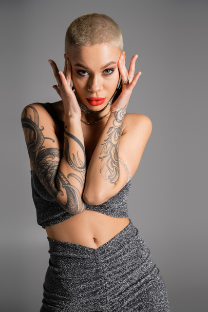 seductive tattooed woman with short hair and bright red lips touching head and looking at camera isolated on grey - Foto, Bild
