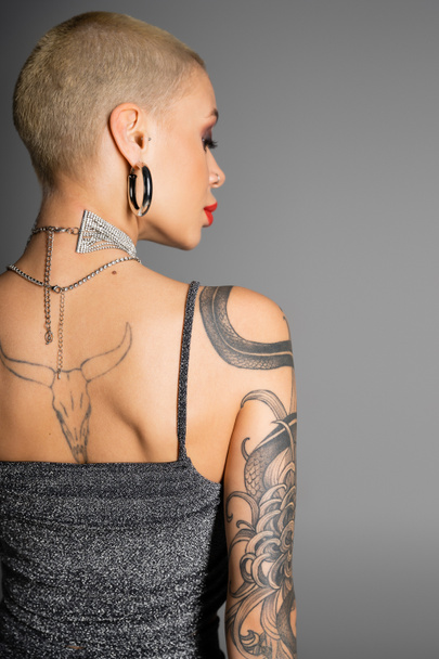 back view of short haired tattooed woman in metal necklaces and shiny top standing isolated on grey - Photo, Image