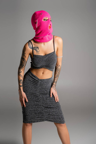 seductive tattooed woman in lurex clothes and pink balaclava looking away isolated on grey - Photo, Image