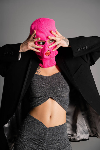 sexy mujer tatuada en ropa elegante cogida de la mano cerca de la cara en pasamontañas rosa mientras mira hacia otro lado aislado en gris - Foto, imagen