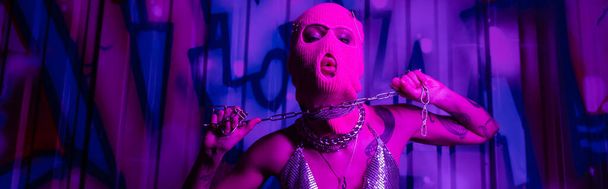 mujer apasionada en pasamontañas posando con cadena de plata cerca de graffiti en luz púrpura, pancarta - Foto, imagen