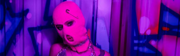 névtelen nő rózsaszín maszkban a kamerát nézi a színes graffiti közelében lila neon fényben, banner - Fotó, kép