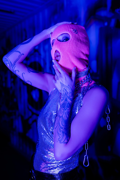 γυναίκα με τατουάζ σε γυαλιστερό τοπ και balaclava κρατώντας τα χέρια κοντά στο πρόσωπο, ενώ κοιτάζοντας κάμερα σε μπλε και μωβ φως - Φωτογραφία, εικόνα