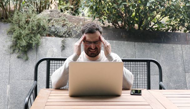 Homem com dor de cabeça usando laptop enquanto sentado ao ar livre no banco. - Foto, Imagem