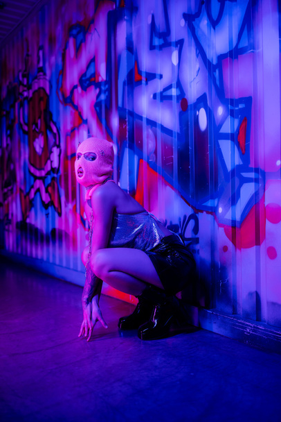 teljes hossza nő elegáns ruhák és símaszk ül fal mellett graffiti kék és lila világítás - Fotó, kép
