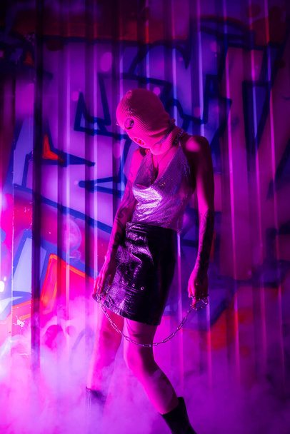 sexy woman in balaclava and shiny top with leather skirt standing with chain near colorful graffiti in purple smoke - Photo, Image