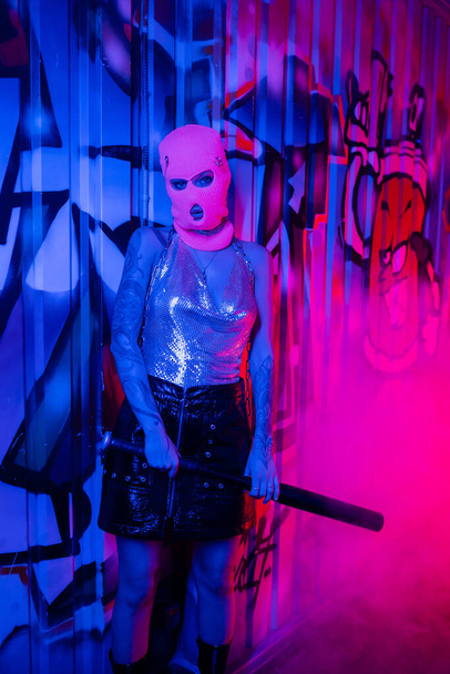 sexy woman in shiny top and balaclava holding baseball bat near colorful graffiti in blue light with pink smoke - Foto, afbeelding