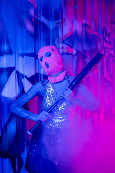 hartstochtelijke vrouw in bivakmuts staande met honkbalknuppel in de buurt van blauwe muur met graffiti in paarse rook - Foto, afbeelding