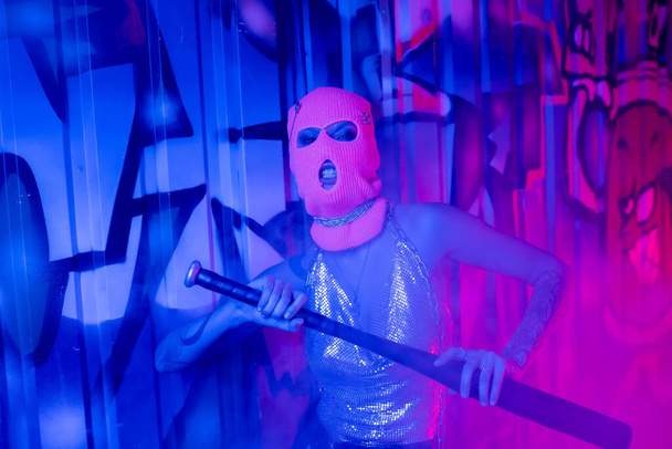 provocative woman in balaclava and silver top grimacing with baseball bat near graffiti in blue and purple light - Photo, Image