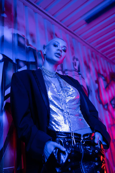 vue à angle bas de la femme sexy en haut métallique et veste noire posant avec les mains dans les poches près des graffitis en lumière bleue et violette - Photo, image