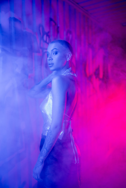 seductive short haired woman in silver top looking at camera near wall with graffiti in blue and pink light with smoke - Photo, Image