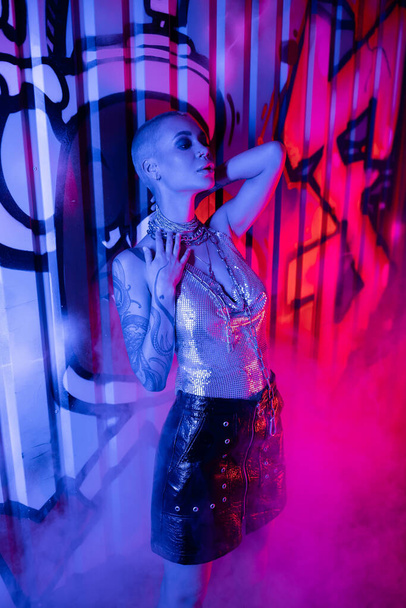 passionate tattooed woman in metallic top and black leather skirt standing near colorful graffiti in blue and pink light with smoke - Photo, Image