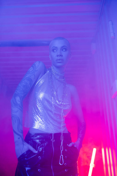 sexy woman in metallic top standing with hands in pockets of the black leather skirt in blue and pink foggy lighting - Fotó, kép