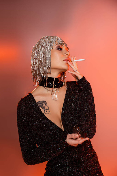 tattooed woman in shiny wig and black sexy dress smoking cigarette on pink and orange background - Φωτογραφία, εικόνα