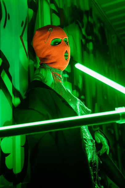 sexy woman in orange balaclava and black blazer looking at camera near bright neon lamps and green wall with graffiti - Photo, Image
