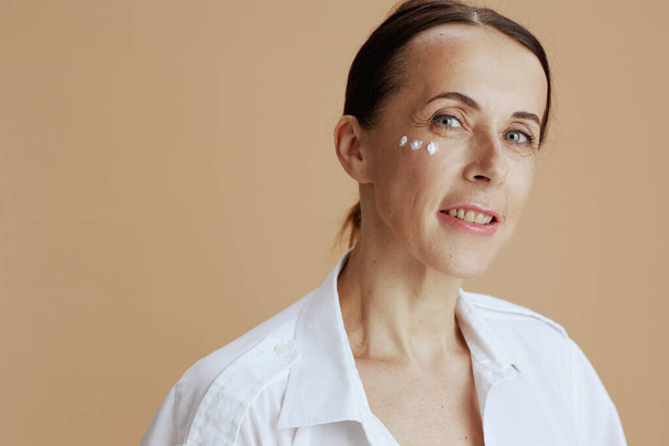Portrait of happy modern 40 years old woman with eye cream on face in white shirt against beige background. - Foto, Bild