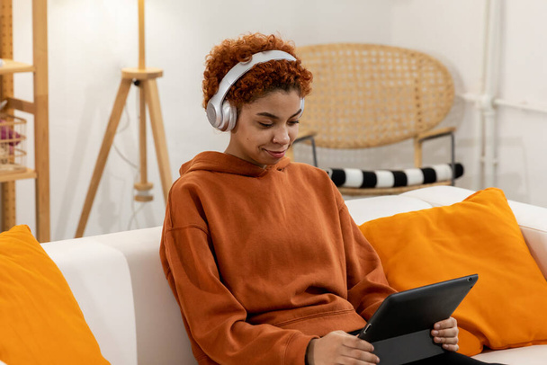 Прекрасна афро-американська дівчина з афроамериканською зачіскою в навушниках посміхається слухати музику вдома. Молода жінка любить слухати улюблену музику на планшеті. Люди ведуть радісний спосіб життя - Фото, зображення
