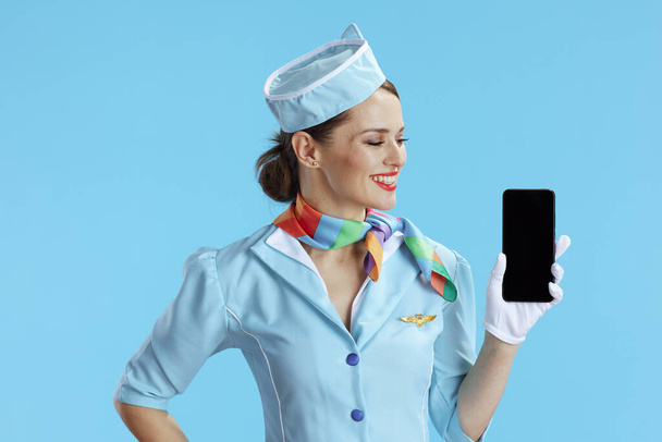 smiling elegant flight attendant woman on blue background in blue uniform showing smartphone blank screen. - Foto, immagini