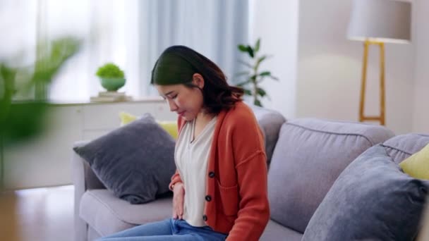 Pain, sick and a woman feeling stomach for ibs, problem and insecurity. Sad, depression and Asian girl touching her belly for body dysmorphia, illness and uncomfortable gas on the lounge sofa. - Footage, Video