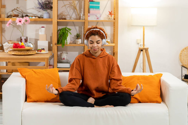 Yoga Achtsamkeitsmeditation. Junge gesunde Afrikanerin praktiziert Yoga zu Hause. Frau sitzt in Lotus-Pose auf Couch meditiert lächelnd entspannt drinnen. Mädchen machen Atemübungen. Yoga zu Hause - Foto, Bild