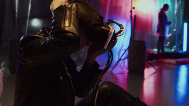 Вид сбоку на молодую кавказскую женщину с бритой головой в кожаном костюме, надевающую очки VR, сидящую в студии с пластиковой пленкой на стенах и неоновыми огнями - Кадры, видео