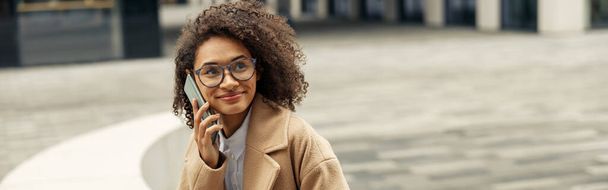 Positive afro american woman wearing glasses talking phone with friends during city walking - 写真・画像