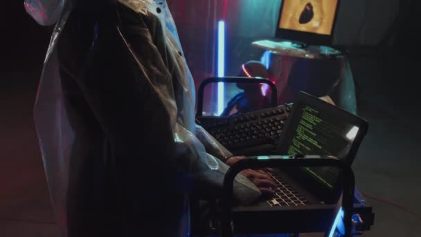 Наклонение вверх боковой вид молодой женщины в плаще, кодирование на портативный компьютер, стоя в убежище с пластиковой пленкой на стенах и неоновых огней - Кадры, видео