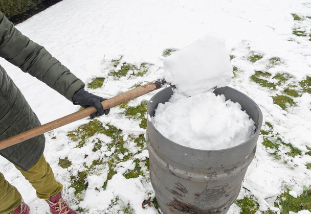 una mujer de mediana edad está recogiendo nieve en un barril con una pala. Para regar aún más las plantas en un invernadero, el concepto de proteger el medio ambiente y conservar los recursos naturales - Foto, Imagen