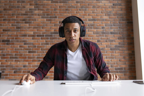 joven jugador afroamericano juega videojuego en el ordenador en casa, hombre en auriculares se sienta en la mesa y mira el monitor - Foto, imagen