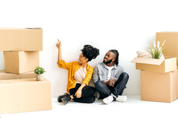 Yeni bir dairenin iç tasarımını planlıyorum. Neşeli Afrikalı Amerikalı evli çift yerde, ellerinde ev eşyalarıyla karton kutuların arasında ilk evlerinde tamirat yapmayı planlıyorlar. - Fotoğraf, Görsel