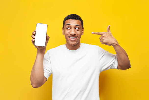 young african american guy in white t-shirt shows blank screen of smartphone on yellow isolated background, man holds phone and advertises screen for mock-up - Photo, Image