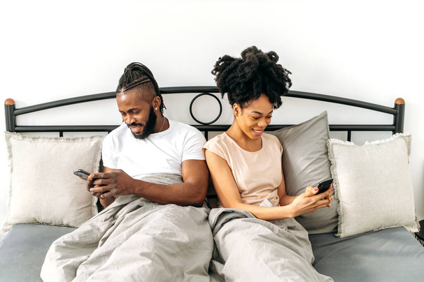 Positivo pareja afroamericana casada, cónyuges felices, esposa y marido se sientan en una cama acogedora en el dormitorio, utilizan sus teléfonos inteligentes, navegar por las redes sociales, noticias, mensajes con amigos, sonrisa - Foto, imagen