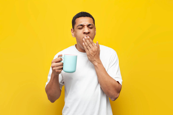 junger müder afrikanisch-amerikanischer Typ in weißem T-Shirt hält eine Tasse Kaffee in der Hand und gähnt auf gelbem isolierten Hintergrund, Mann mit Getränk bekommt nicht genug Schlaf, das Konzept der Schläfrigkeit und Schlaflosigkeit - Foto, Bild