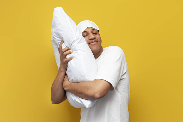 young african american guy in pajamas and sleep mask hugs soft comfortable pillow and smiles on yellow isolated background, the man sleeps and sees good pleasant dreams - Fotografie, Obrázek