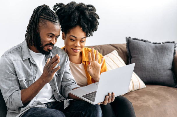 Positiva pareja afroamericana sentada en un sofá en casa en la sala de estar, hablando en videoconferencia con amigos o familiares por computadora portátil, hacer un gesto de saludo, sonreír. Comunicación de vídeo en línea - Foto, Imagen