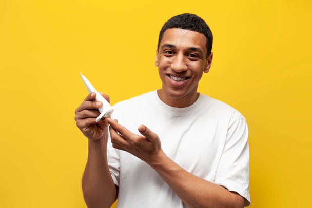 young african american guy in white t-shirt squeezes out cream from tube on yellow isolated background, man uses facial cosmetics - Photo, Image